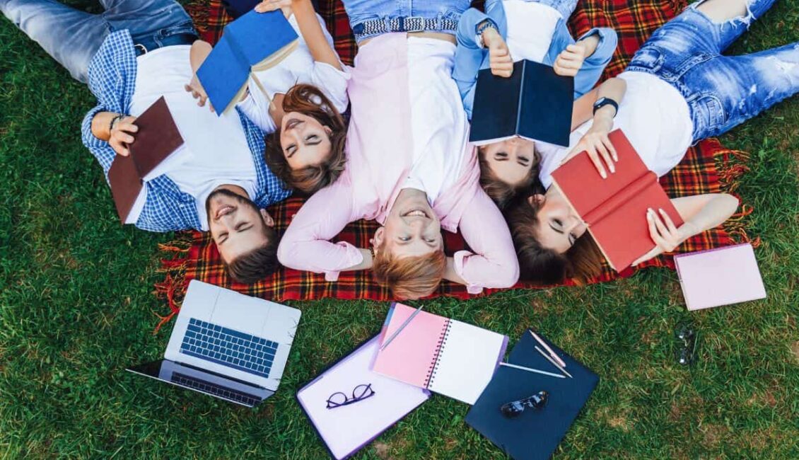 Öğrenciler çimenlik alanda sırt üstü yatarak kitap okumaktalar. Yanlarında laptop gözlük, kalemler ve defterler bulunmakta.